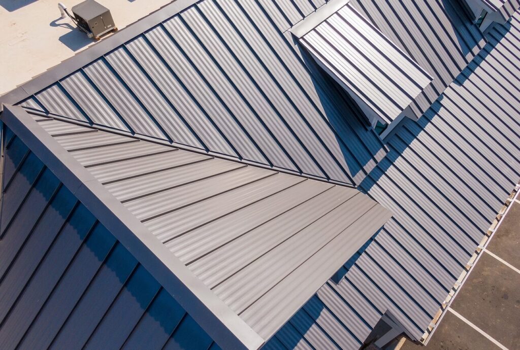 Aerial view of a commercial building with light gray standing seam metal roof panels showing multiple roof planes, ridges, and valleys meeting at various angles