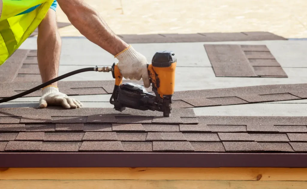Roofing worker using an orange pneumatic nail gun to fasten brown asphalt shingles along a roof edge